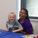 Two young girls are seated together at a table covered with a blue tablecloth, both smiling and laughing joyfully. The older girl, wearing glasses and a purple shirt, has her arm around the younger girl who is wearing a teal jersey-style shirt. The setting appears to be an indoor gathering or party with food visible on the table.