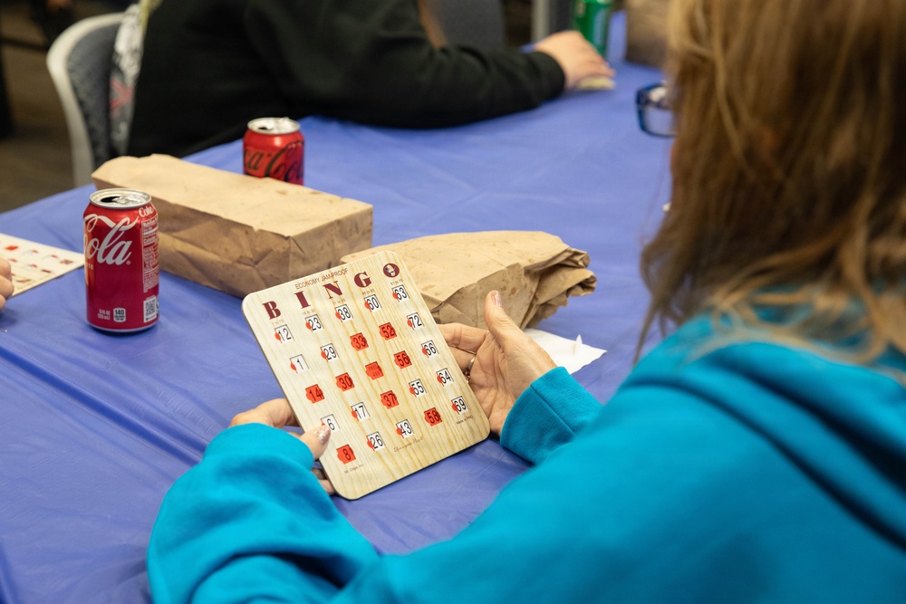 A person wearing a teal/turquoise sweatshirt is holding a wooden bingo card at a table covered with a blue tablecloth. The setting appears to be a casual bingo game event, with multiple players seated at the table. Brown paper bags and Coca-Cola cans are visible on the table, suggesting refreshments or prizes are part of the event.