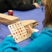 A person wearing a teal/turquoise sweatshirt is holding a wooden bingo card at a table covered with a blue tablecloth. The setting appears to be a casual bingo game event, with multiple players seated at the table. Brown paper bags and Coca-Cola cans are visible on the table, suggesting refreshments or prizes are part of the event.