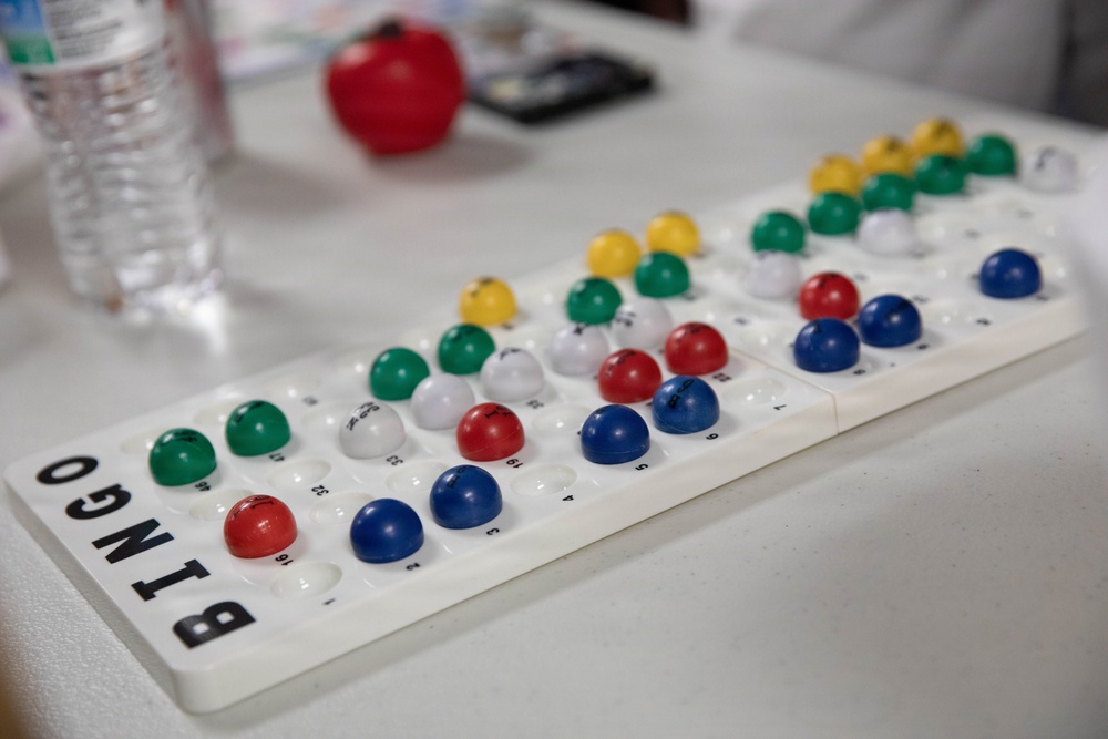 A white plastic Bingo ball tracking board is displayed on a white table, with colored dome-shaped markers inserted into numbered slots indicating called numbers. The board features the word 'BINGO' printed in bold black letters along its side. Various colored markers — blue, red, green, yellow, and white — are placed across the board to track which numbers have been called during a game.