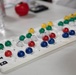 A white plastic Bingo ball tracking board is displayed on a white table, with colored dome-shaped markers inserted into numbered slots indicating called numbers. The board features the word 'BINGO' printed in bold black letters along its side. Various colored markers — blue, red, green, yellow, and white — are placed across the board to track which numbers have been called during a game.
