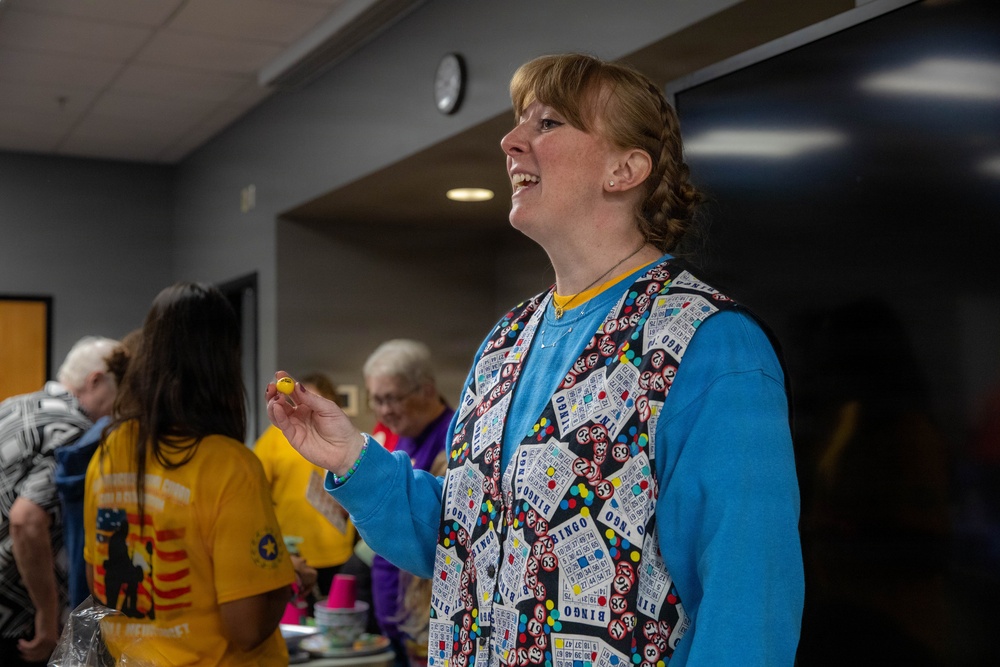 A woman stands in the foreground of what appears to be a community bingo event, smiling and holding a small bingo ball. She is wearing a bingo-themed vest over a blue sweatshirt, suggesting she may be a caller or organizer. Several other people are visible in the background in a community hall or recreation room setting.