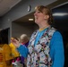 A woman stands in the foreground of what appears to be a community bingo event, smiling and holding a small bingo ball. She is wearing a bingo-themed vest over a blue sweatshirt, suggesting she may be a caller or organizer. Several other people are visible in the background in a community hall or recreation room setting.