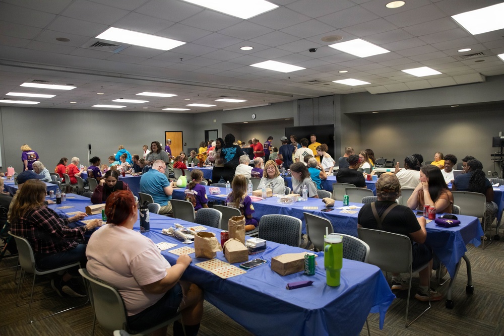 A large indoor gathering is taking place in what appears to be a community center or institutional meeting room. Numerous people of various ages are seated at long rectangular tables covered with blue tablecloths, engaged in what appears to be a bingo game or similar activity. The event has a casual, social atmosphere with food and beverages visible on the tables.