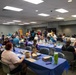 A large indoor gathering is taking place in what appears to be a community center or institutional meeting room. Numerous people of various ages are seated at long rectangular tables covered with blue tablecloths, engaged in what appears to be a bingo game or similar activity. The event has a casual, social atmosphere with food and beverages visible on the tables.