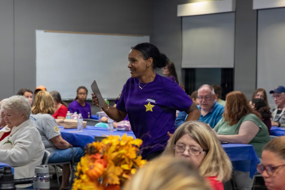 A woman wearing a purple t-shirt with a gold star emblem stands among seated attendees in what appears to be a community gathering or event in a meeting hall. She is holding papers and appears to be speaking or presenting to the group. Tables are covered with blue tablecloths and decorated with autumn floral arrangements, suggesting a fall-themed community event.