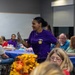 A woman wearing a purple t-shirt with a gold star emblem stands among seated attendees in what appears to be a community gathering or event in a meeting hall. She is holding papers and appears to be speaking or presenting to the group. Tables are covered with blue tablecloths and decorated with autumn floral arrangements, suggesting a fall-themed community event.