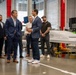 A group of professionals are touring what appears to be an aerospace or defense manufacturing facility. Several individuals in business attire are engaged in conversation near a large white aircraft or drone component on a wheeled cart. The setting is an industrial hangar or production floor with high ceilings and red structural columns.