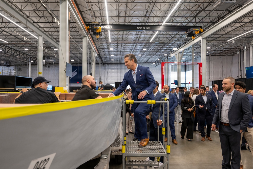 A man in a navy blue suit stands elevated on a metal step platform inside a large industrial manufacturing facility, engaging with workers near what appears to be a boat or vessel hull under construction. He is surrounded by a group of approximately 15-20 people in business attire, suggesting an official tour or facility visit. The facility features overhead crane systems rated at 10 tons and high-bay industrial lighting.