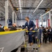 A man in a navy blue suit stands elevated on a metal step platform inside a large industrial manufacturing facility, engaging with workers near what appears to be a boat or vessel hull under construction. He is surrounded by a group of approximately 15-20 people in business attire, suggesting an official tour or facility visit. The facility features overhead crane systems rated at 10 tons and high-bay industrial lighting.