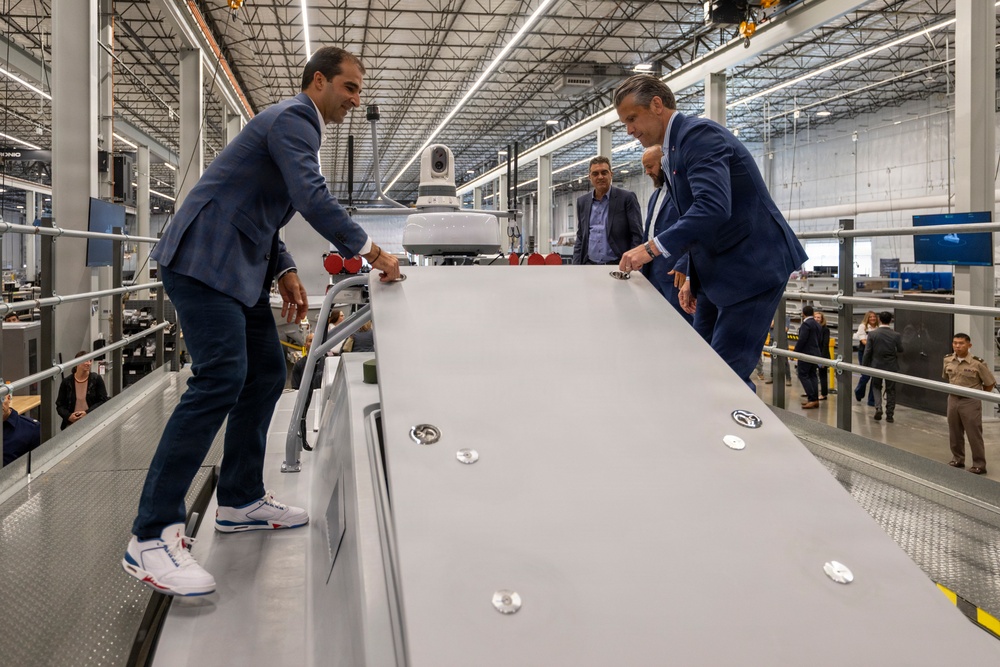 Several individuals in business attire are examining a large gray industrial panel or surface inside a modern manufacturing facility. Two men in blue suits lean over the panel from opposite sides, appearing to inspect or demonstrate its features. The setting is a large, well-lit industrial warehouse with visible machinery and equipment in the background.