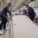 Several individuals in business attire are examining a large gray industrial panel or surface inside a modern manufacturing facility. Two men in blue suits lean over the panel from opposite sides, appearing to inspect or demonstrate its features. The setting is a large, well-lit industrial warehouse with visible machinery and equipment in the background.