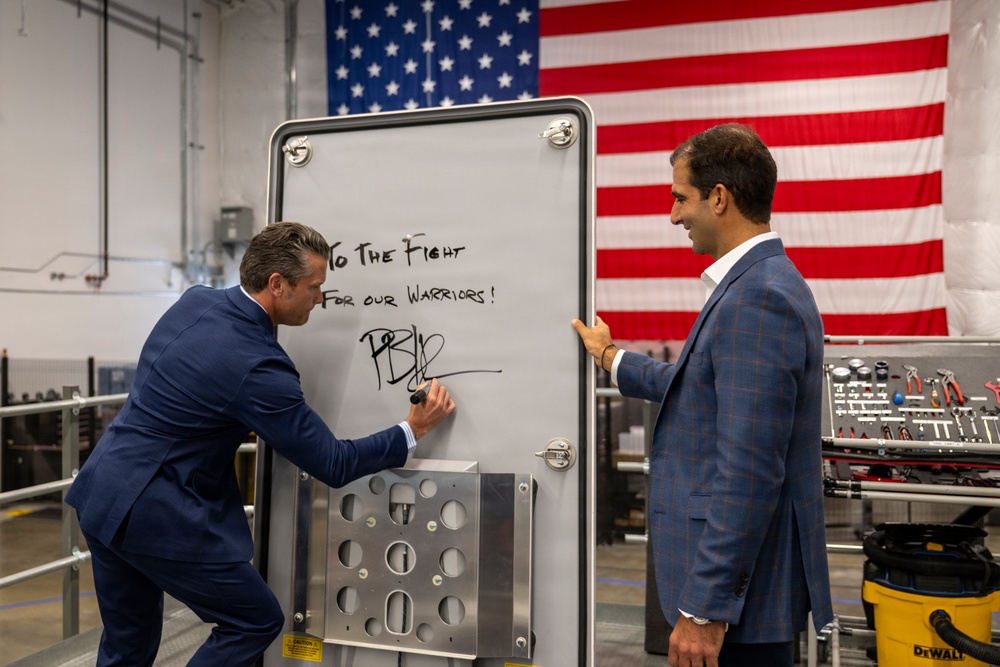 Two men in business suits are in an industrial or manufacturing facility, where one is writing a message on a whiteboard-style panel. The written message reads 'To The Fight For our Warriors!' followed by a signature. A large American flag is displayed prominently in the background.