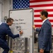 Two men in business suits are in an industrial or manufacturing facility, where one is writing a message on a whiteboard-style panel. The written message reads 'To The Fight For our Warriors!' followed by a signature. A large American flag is displayed prominently in the background.