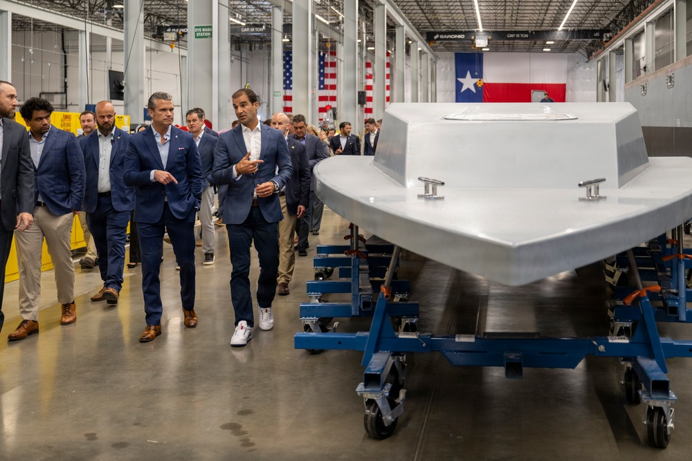 A group of professionally dressed individuals, primarily in navy suits, are touring what appears to be a manufacturing or assembly facility. They are walking alongside a large gray vessel or watercraft hull mounted on blue wheeled industrial dollies. An American flag and Texas state flag are visible in the background, suggesting this facility is located in Texas.