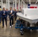 A group of professionally dressed individuals, primarily in navy suits, are touring what appears to be a manufacturing or assembly facility. They are walking alongside a large gray vessel or watercraft hull mounted on blue wheeled industrial dollies. An American flag and Texas state flag are visible in the background, suggesting this facility is located in Texas.