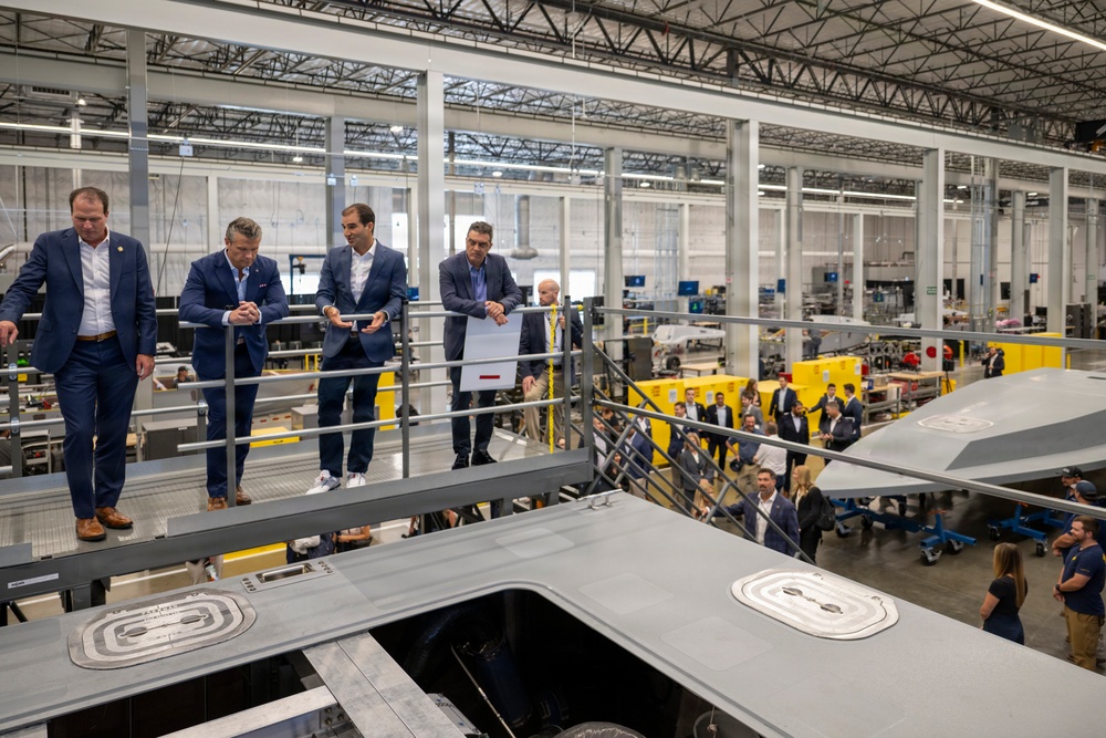 A group of formally dressed individuals are touring what appears to be an aerospace or defense manufacturing facility. They are standing on an elevated mezzanine level overlooking a large production floor, with what appears to be a gray stealth aircraft or drone visible in the foreground. The facility is modern, well-lit, and contains yellow industrial equipment and multiple workstations.