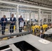 A group of formally dressed individuals are touring what appears to be an aerospace or defense manufacturing facility. They are standing on an elevated mezzanine level overlooking a large production floor, with what appears to be a gray stealth aircraft or drone visible in the foreground. The facility is modern, well-lit, and contains yellow industrial equipment and multiple workstations.
