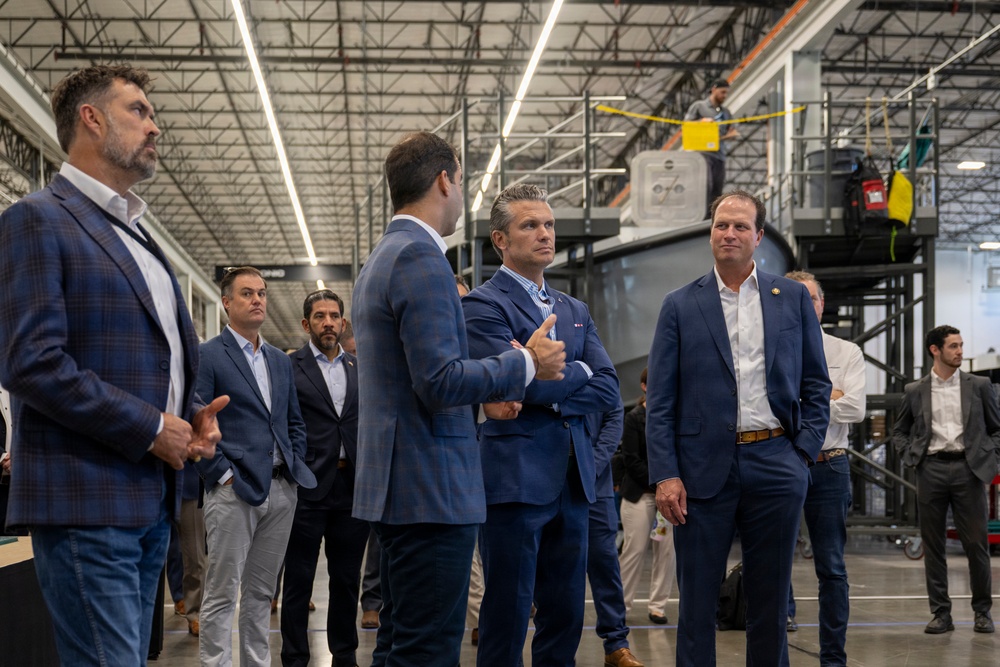 A group of professionally dressed individuals, primarily in business suits, are gathered inside a large industrial facility or manufacturing warehouse. The setting appears to be a formal tour or briefing, with several men engaged in conversation in the foreground while others observe in the background. Large industrial equipment and elevated platforms are visible in the facility.