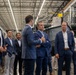 A group of professionally dressed individuals, primarily in business suits, are gathered inside a large industrial facility or manufacturing warehouse. The setting appears to be a formal tour or briefing, with several men engaged in conversation in the foreground while others observe in the background. Large industrial equipment and elevated platforms are visible in the facility.