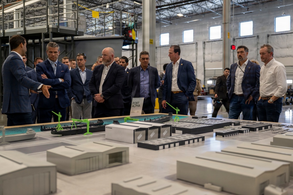 A group of approximately 15-20 professionals, mostly dressed in business attire, are gathered around a large architectural scale model inside an industrial warehouse or facility. The model appears to represent a port development project labeled 'Port Alpha,' featuring miniature buildings, cranes, and waterway elements. The setting suggests a formal presentation or site briefing for stakeholders or officials.