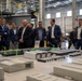 A group of approximately 15-20 professionals, mostly dressed in business attire, are gathered around a large architectural scale model inside an industrial warehouse or facility. The model appears to represent a port development project labeled 'Port Alpha,' featuring miniature buildings, cranes, and waterway elements. The setting suggests a formal presentation or site briefing for stakeholders or officials.