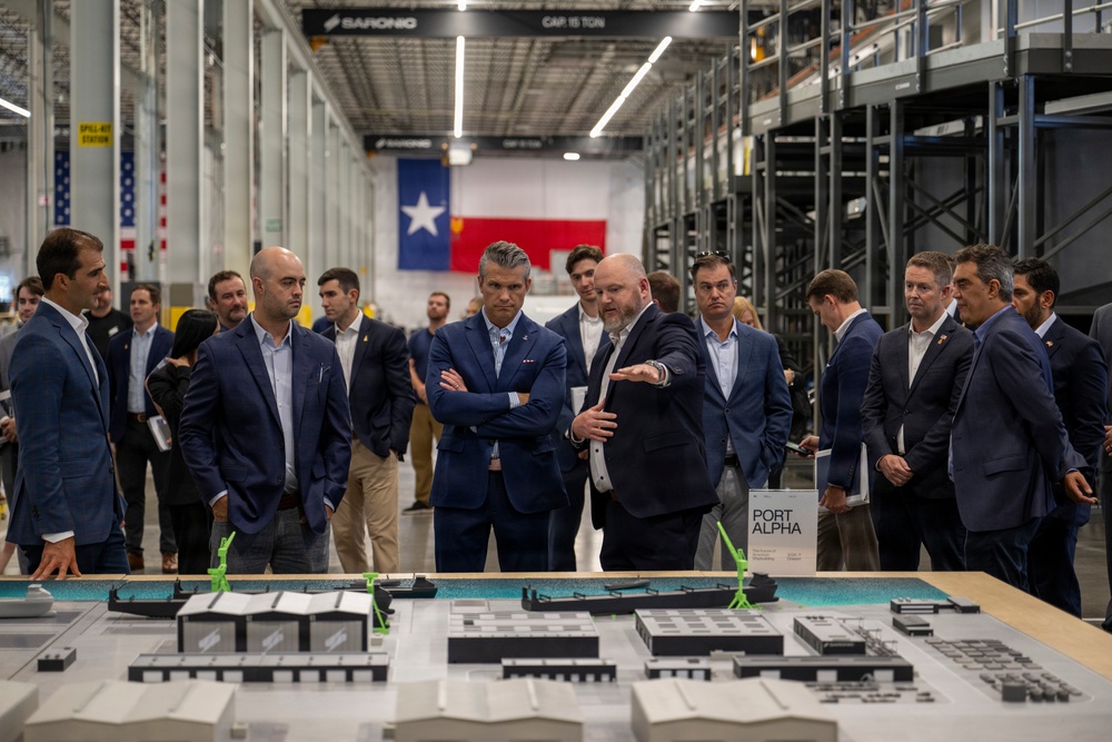 A group of professionally dressed individuals, mostly in dark suits, are gathered around a detailed architectural scale model labeled 'Port Alpha' inside a large industrial facility. One person appears to be presenting or explaining the model to the group. The setting features a Texas state flag and an American flag hanging in the background, along with industrial shelving and overhead cranes.