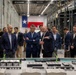 A group of professionally dressed individuals, mostly in dark suits, are gathered around a detailed architectural scale model labeled 'Port Alpha' inside a large industrial facility. One person appears to be presenting or explaining the model to the group. The setting features a Texas state flag and an American flag hanging in the background, along with industrial shelving and overhead cranes.