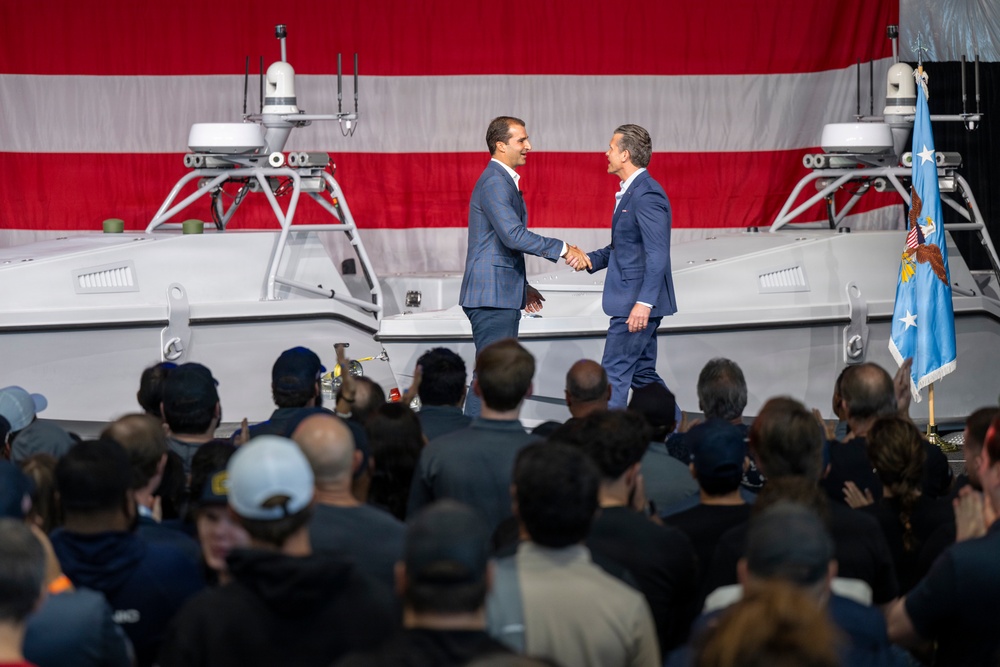 Two men in dark business suits are shaking hands on a stage in front of a large American flag backdrop and what appears to be an unmanned surface vessel (USV) or autonomous military watercraft. A crowd of attendees watches from below the stage, and a light blue flag with an eagle emblem is visible on the right side. The setting suggests a formal ceremony, likely a military or defense industry event.