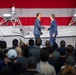 Two men in dark business suits are shaking hands on a stage in front of a large American flag backdrop and what appears to be an unmanned surface vessel (USV) or autonomous military watercraft. A crowd of attendees watches from below the stage, and a light blue flag with an eagle emblem is visible on the right side. The setting suggests a formal ceremony, likely a military or defense industry event.