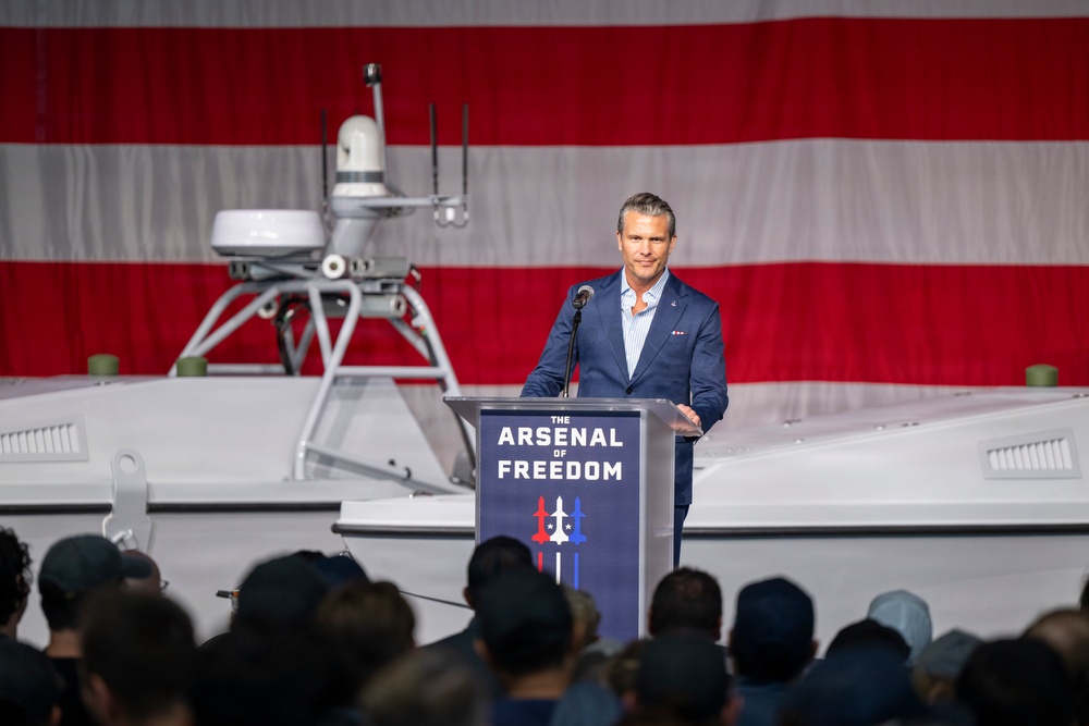 A man in a navy blue suit is speaking at a podium labeled 'The Arsenal of Freedom' in front of a large American flag backdrop. Behind him is what appears to be an unmanned surface vessel (USV) or autonomous naval craft with sensor equipment mounted on top. The event appears to be a formal defense or military technology presentation with a large audience in attendance.