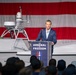 A man in a navy blue suit is speaking at a podium labeled 'The Arsenal of Freedom' in front of a large American flag backdrop. Behind him is what appears to be an unmanned surface vessel (USV) or autonomous naval craft with sensor equipment mounted on top. The event appears to be a formal defense or military technology presentation with a large audience in attendance.