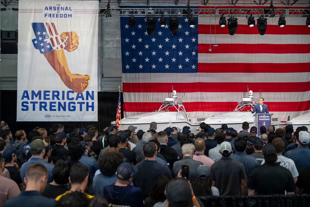 A large indoor event is taking place in what appears to be a hangar or industrial facility, organized under the 'Arsenal of Freedom' initiative by the U.S. Department of War. A speaker addresses a large crowd from a podium in front of a massive American flag backdrop, with what appear to be unmanned surface vessels (USVs) or autonomous watercraft displayed on stage. The event is themed around 'American Strength' as indicated by prominent signage.