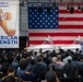 A large indoor event is taking place in what appears to be a hangar or industrial facility, organized under the 'Arsenal of Freedom' initiative by the U.S. Department of War. A speaker addresses a large crowd from a podium in front of a massive American flag backdrop, with what appear to be unmanned surface vessels (USVs) or autonomous watercraft displayed on stage. The event is themed around 'American Strength' as indicated by prominent signage.