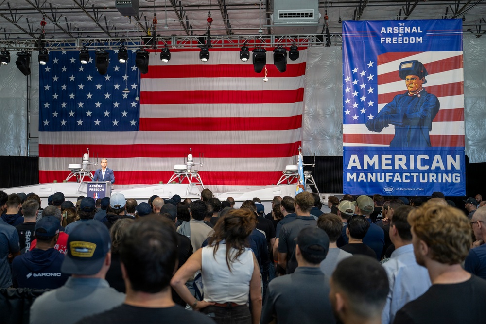 A large indoor event is taking place in what appears to be a manufacturing facility or hangar, with a speaker at a podium addressing a crowd of attendees. The stage is decorated with a large American flag backdrop and features what appear to be unmanned surface vessels or military equipment on display. A prominent banner reading 'The Arsenal of Freedom - American Manufacturing' with a U.S. Department of War logo is displayed on the right side.