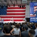 A large indoor event is taking place in what appears to be a manufacturing facility or hangar, with a speaker at a podium addressing a crowd of attendees. The stage is decorated with a large American flag backdrop and features what appear to be unmanned surface vessels or military equipment on display. A prominent banner reading 'The Arsenal of Freedom - American Manufacturing' with a U.S. Department of War logo is displayed on the right side.