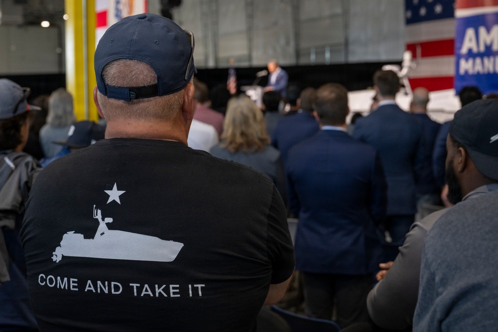 A crowd of attendees is gathered at what appears to be a political or public event held in an industrial or hangar-style venue. The foreground prominently features a man wearing a black t-shirt with a 'Come and Take It' slogan and a boat graphic, along with a navy baseball cap. A speaker is visible at a podium in the background, with American flags and a partial banner reading 'AM[ERICA] MAN[UFACTURING]' or similar text.