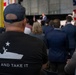 A crowd of attendees is gathered at what appears to be a political or public event held in an industrial or hangar-style venue. The foreground prominently features a man wearing a black t-shirt with a 'Come and Take It' slogan and a boat graphic, along with a navy baseball cap. A speaker is visible at a podium in the background, with American flags and a partial banner reading 'AM[ERICA] MAN[UFACTURING]' or similar text.