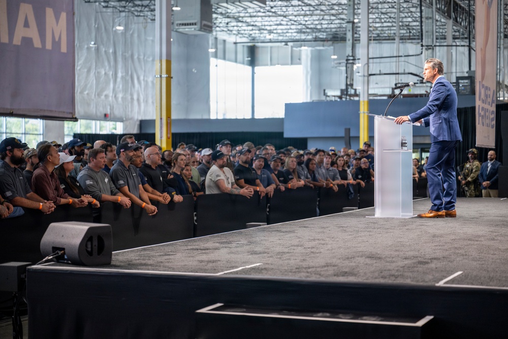 A man in a blue suit is delivering a speech at a podium on a raised stage inside a large industrial or warehouse-style facility. A large crowd of attendees, many wearing casual work attire and orange wristbands, lines the edge of the stage listening attentively. The setting appears to be a formal announcement or company event held within a manufacturing or logistics facility.