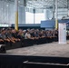 A man in a blue suit is delivering a speech at a podium on a raised stage inside a large industrial or warehouse-style facility. A large crowd of attendees, many wearing casual work attire and orange wristbands, lines the edge of the stage listening attentively. The setting appears to be a formal announcement or company event held within a manufacturing or logistics facility.