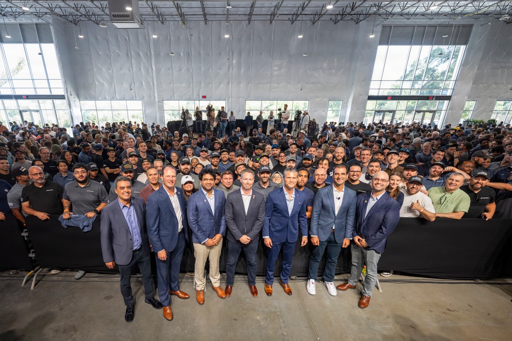 A large group photograph taken inside a modern industrial or event facility with high ceilings and large windows. A row of formally dressed individuals in suits stands at the front, while hundreds of employees or attendees fill the background. The setting and attire suggest a corporate event, company milestone, or facility opening ceremony.