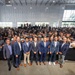 A large group photograph taken inside a modern industrial or event facility with high ceilings and large windows. A row of formally dressed individuals in suits stands at the front, while hundreds of employees or attendees fill the background. The setting and attire suggest a corporate event, company milestone, or facility opening ceremony.