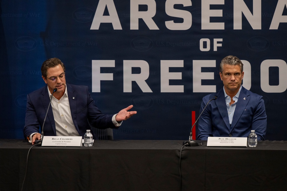 Two men are seated at a panel table in front of a large banner reading 'Arsenal of Freedom' with 'U.S. Department of War' branding visible in the background. Name placards identify the individuals as Bryan Chambers (Co-Founder &amp; President, Capital Factory) on the left and Pete Hegseth (Secretary of War, Department of War) on the right. The man on the left appears to be speaking while gesturing with his hand.
