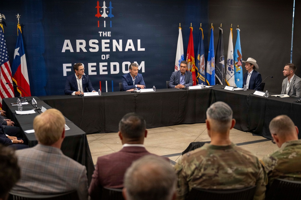 A formal roundtable meeting is taking place at a venue branded 'The Arsenal of Freedom,' featuring several individuals seated at a U-shaped table arrangement. Military personnel in camouflage uniforms and civilians in business attire are present as audience members. The setting appears to be a defense or military-industry conference or briefing.