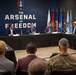 A formal roundtable meeting is taking place at a venue branded 'The Arsenal of Freedom,' featuring several individuals seated at a U-shaped table arrangement. Military personnel in camouflage uniforms and civilians in business attire are present as audience members. The setting appears to be a defense or military-industry conference or briefing.