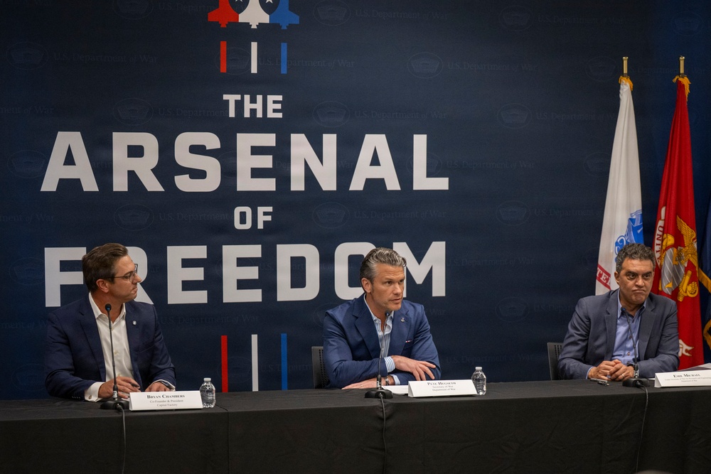 Three men in business attire are seated at a panel table in front of a large navy blue backdrop reading 'The Arsenal of Freedom' with U.S. Department of War branding. Name placards are visible on the table, with the center placard reading 'Pete Hegseth' identified as Department of War, and the left placard reading 'Bryan Chambers' identified as Co-Founder &amp; President of Capitol Forces. A Marine Corps flag and what appears to be a state flag are visible on the right side.