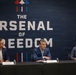 Three men in business attire are seated at a panel table in front of a large navy blue backdrop reading 'The Arsenal of Freedom' with U.S. Department of War branding. Name placards are visible on the table, with the center placard reading 'Pete Hegseth' identified as Department of War, and the left placard reading 'Bryan Chambers' identified as Co-Founder &amp; President of Capitol Forces. A Marine Corps flag and what appears to be a state flag are visible on the right side.