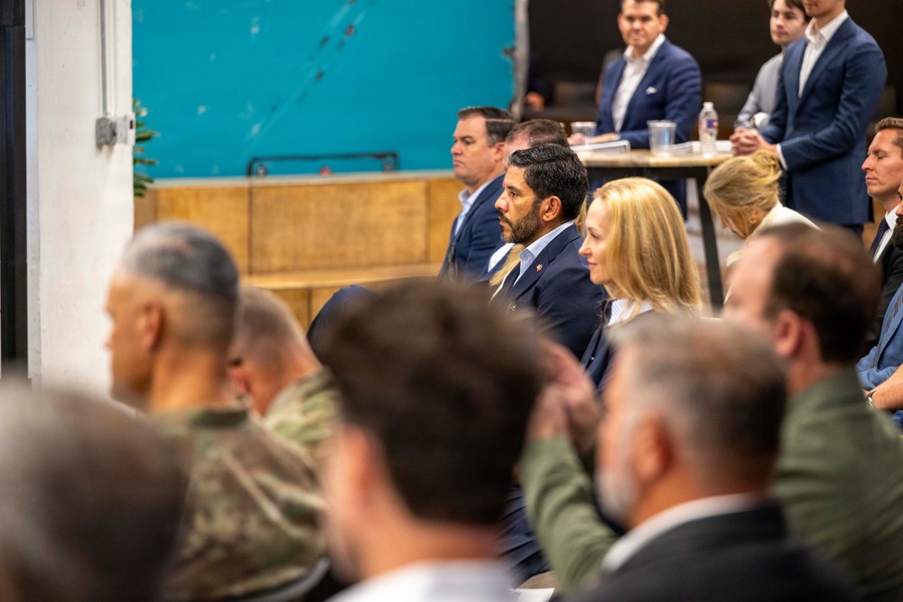 A formal gathering or briefing is taking place in what appears to be an industrial or converted event space. The audience includes a mix of civilians in business attire and at least one individual in military camouflage uniform, suggesting a civil-military or government-related meeting. Attendees are seated and appear to be listening to a presentation or speaker out of frame.