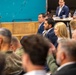 A formal gathering or briefing is taking place in what appears to be an industrial or converted event space. The audience includes a mix of civilians in business attire and at least one individual in military camouflage uniform, suggesting a civil-military or government-related meeting. Attendees are seated and appear to be listening to a presentation or speaker out of frame.