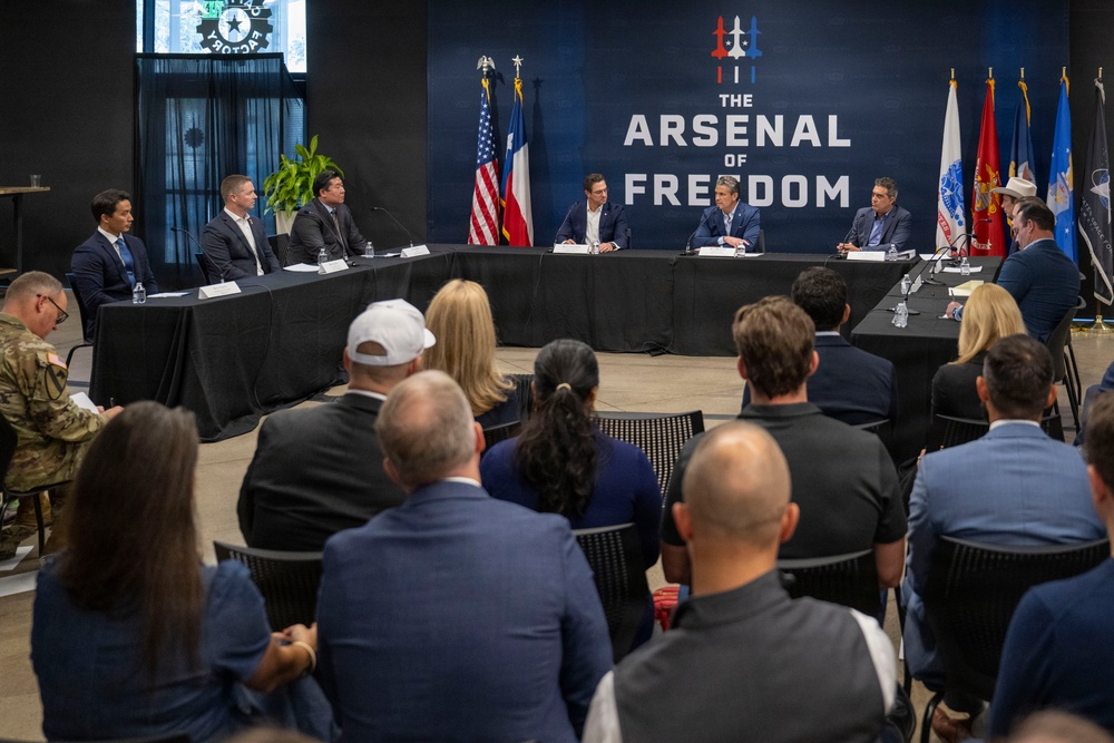 A formal roundtable meeting is taking place at a venue branded 'The Arsenal of Freedom,' with several officials and attendees seated at a curved conference table. The setting includes American and Texas state flags, along with multiple military branch flags displayed in the background. An audience of approximately 20-25 people is seated facing the panel.